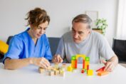 A caregiver in blue scrubs is sitting at a table with an elderly man in a gray t-shirt. They are focused on playing with building blocks, which appear to be part of a cognitive or occupational therapy exercise. A colorful archway structure is built from the blocks, and other wooden blocks are scattered on the white table. They are indoors, with a couch and a potted plant visible in the blurred background.