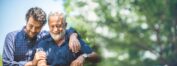 A candid outdoor photo features an adult son with his senior father. The son, wearing a plaid shirt, has his arm around his father's shoulder. The father, with a white beard and a dark blue collared shirt, is smiling with his eyes closed. Both men appear happy and relaxed. The background is a bright, softly blurred green and white of trees and sky.