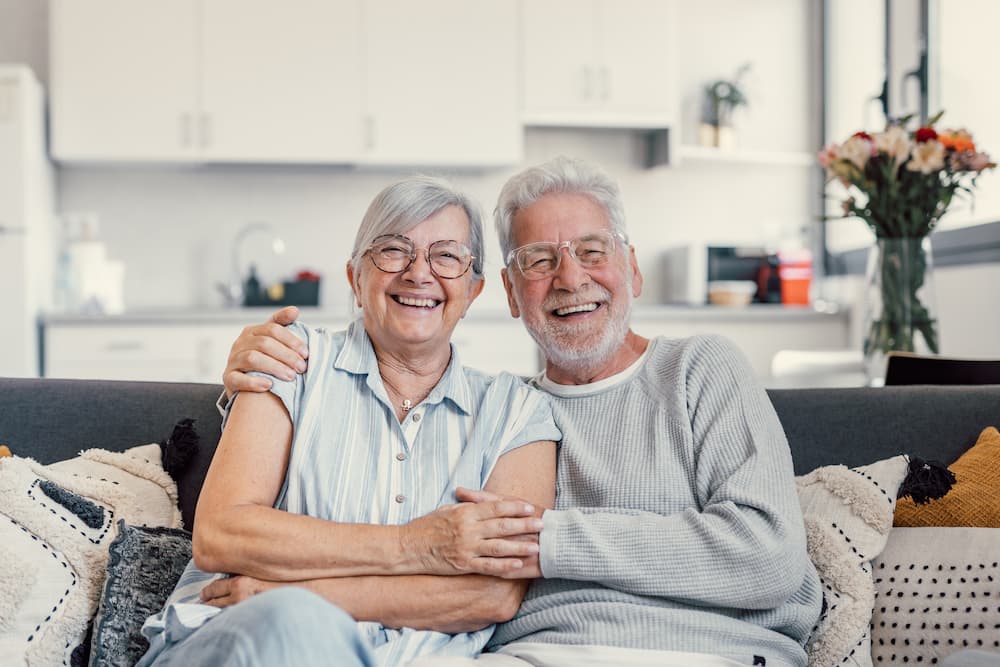 Happy elderly couple sitting on a couch in their modern, bright home, illustrating the importance of regular medical care and personal maintenance for a healthy and fulfilling retirement.