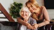 A candid indoor photograph of a happy mother and adult daughter sharing an embrace. The elderly mother with short white hair is seated and smiling directly at the camera. The daughter, with long blonde hair and wearing a tan top, is standing behind her mother, leaning in with her arms around her mother's shoulders and smiling with her eyes closed. They are seated on a dark couch, with a blurred interior background featuring a houseplant and a wooden staircase.