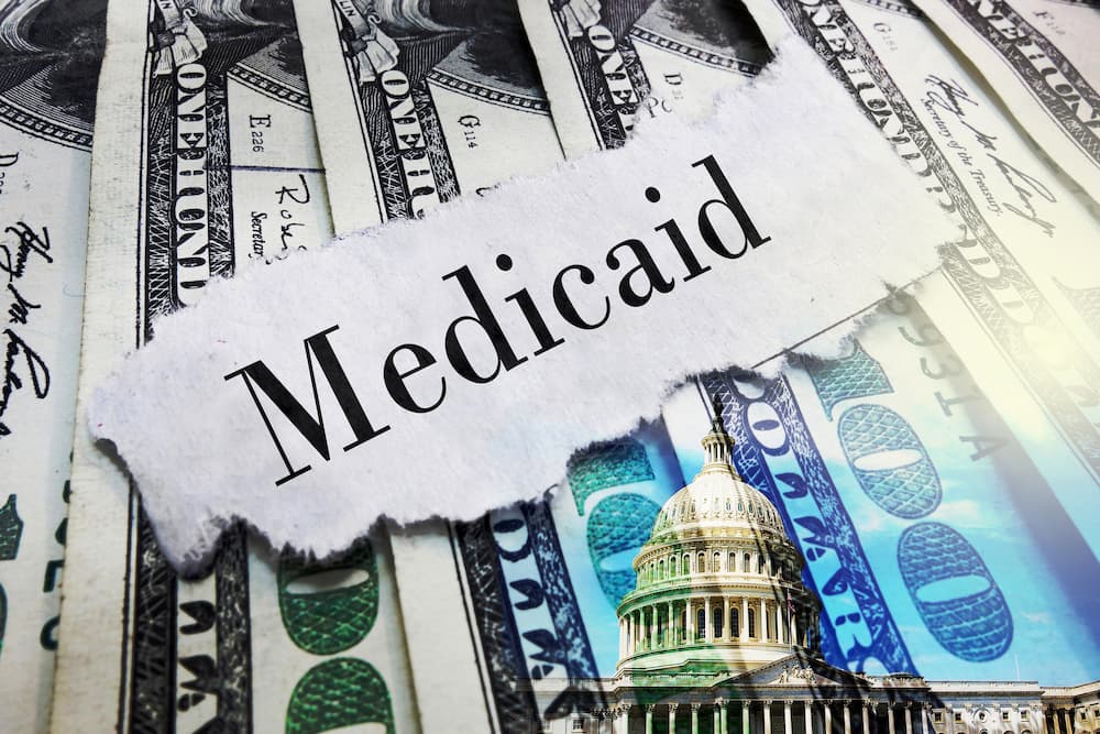 Torn paper with the word "Medicaid" on top of US dollar bills and a double exposure image of the US Capitol Building, emphasizing the need to maintain eligibility and coverage for government healthcare programs.