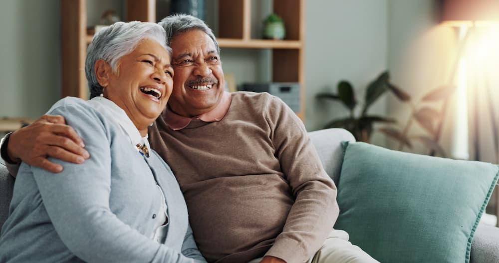 Happy, older couple embracing and laughing on a couch, illustrating the joyful well-being and consistent personal care provided by senior care for veterans.