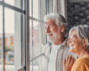 Happy, smiling senior couple standing together and looking optimistically out a large window in their home.