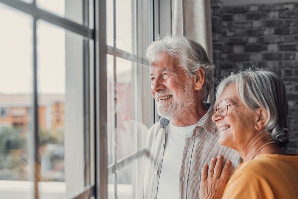 Happy, smiling senior couple standing together and looking optimistically out a large window in their home.