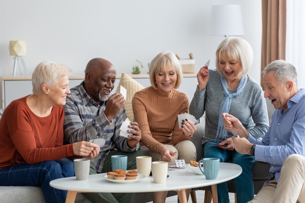 Diverse seniors sitting together on a couch, laughing and playing a card game, highlighting social activities in a senior living community.